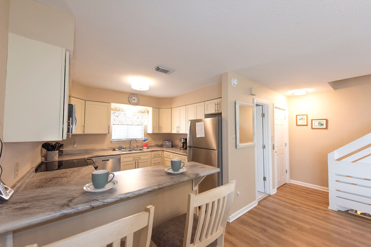 Cook a full vacation meal without leaving anything behind &mdash; the kitchen has a proper breakfast bar, white cabinets stocked with what you need, and three barstools for whoever wanders in first.