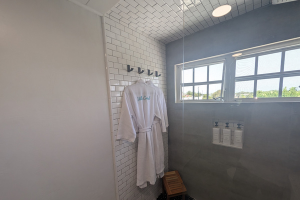 Walk-in shower at Villa Coral featuring white subway tile, large gray porcelain panels, dark pebble flooring, a teak shower stool, and embroidered luxury spa robes.
