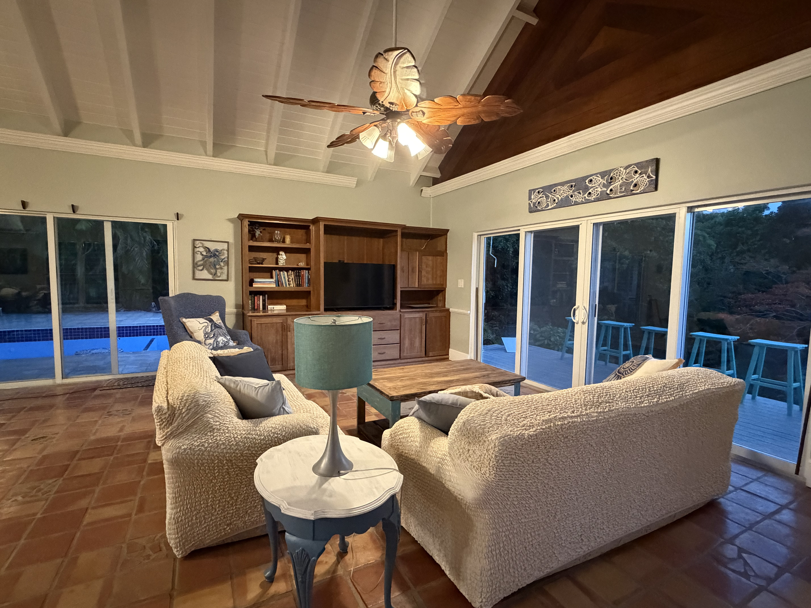 Sliding glass doors surround the room, flooding the space with natural light and opening directly to a private pool deck on one side and a deck with outdoor seating on the other—bringing seamless indoor-outdoor living right to your fingertips.