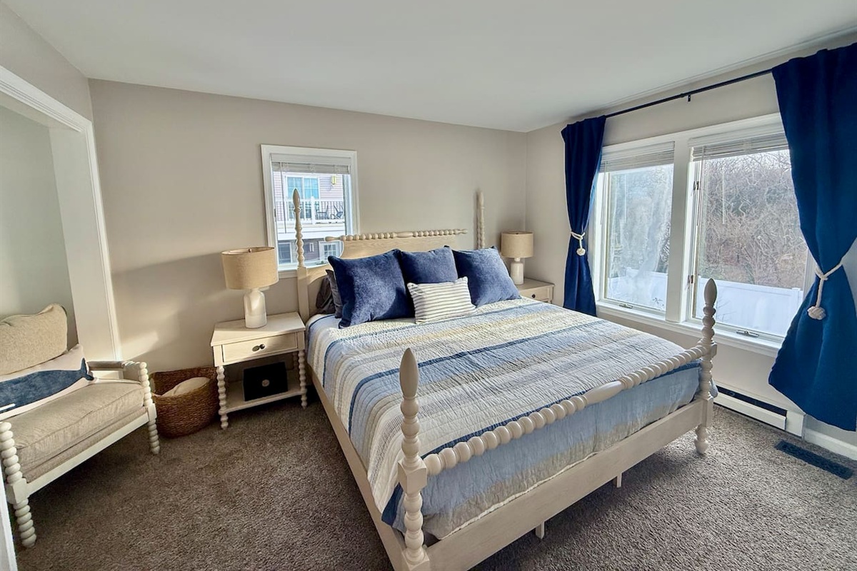This bright bedroom features water views with a white spindle bed with blue-striped bedding, a cozy armchair with a whale pillow, and deep blue curtains, creating a tranquil, well-coordinated seaside retreat for guests to relax in comfort.