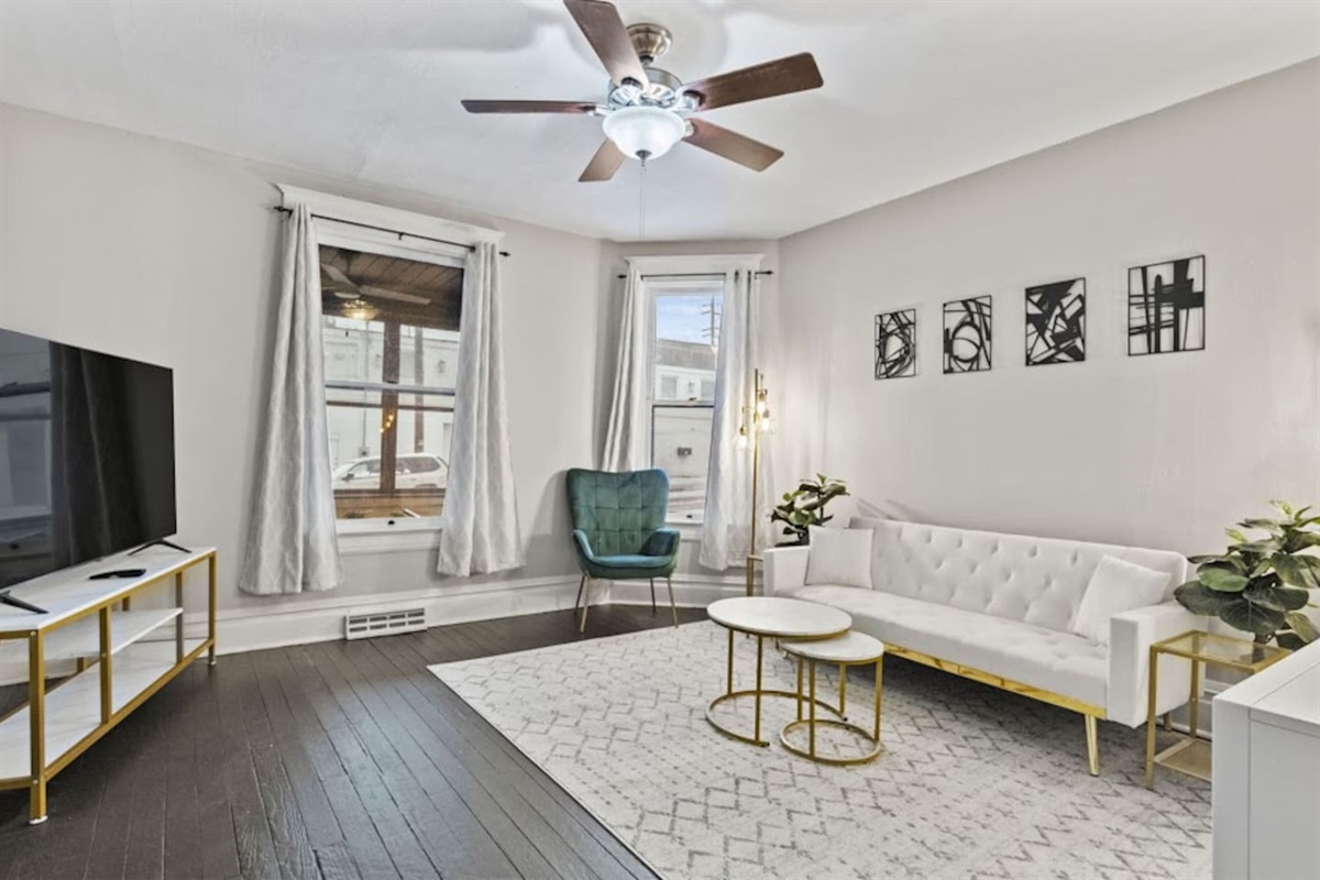 Bright living room featuring a large window with stylish curtains, a chandelier ceiling fan, an elegant sofa, tasteful decorations, mini side tables, and a polished, sophisticated floor design—perfectly blending comfort and modern elegance