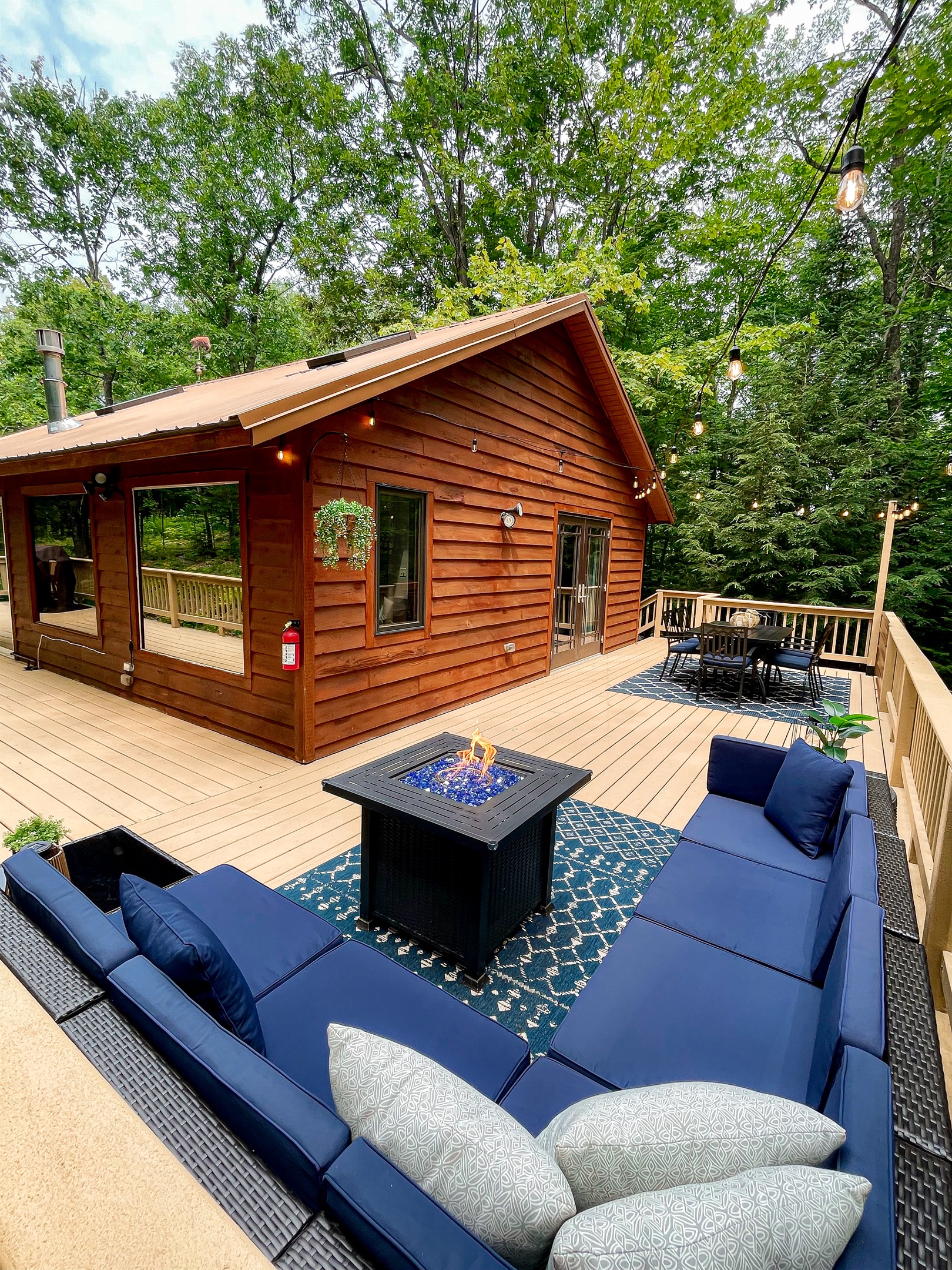 Back deck dining table & outdoor lounge couch, propane fireplace, lit by string lights. Views surrounded by the woods. Outdoor amenities Seasonal (3rd week of May - 3rd week of October) 