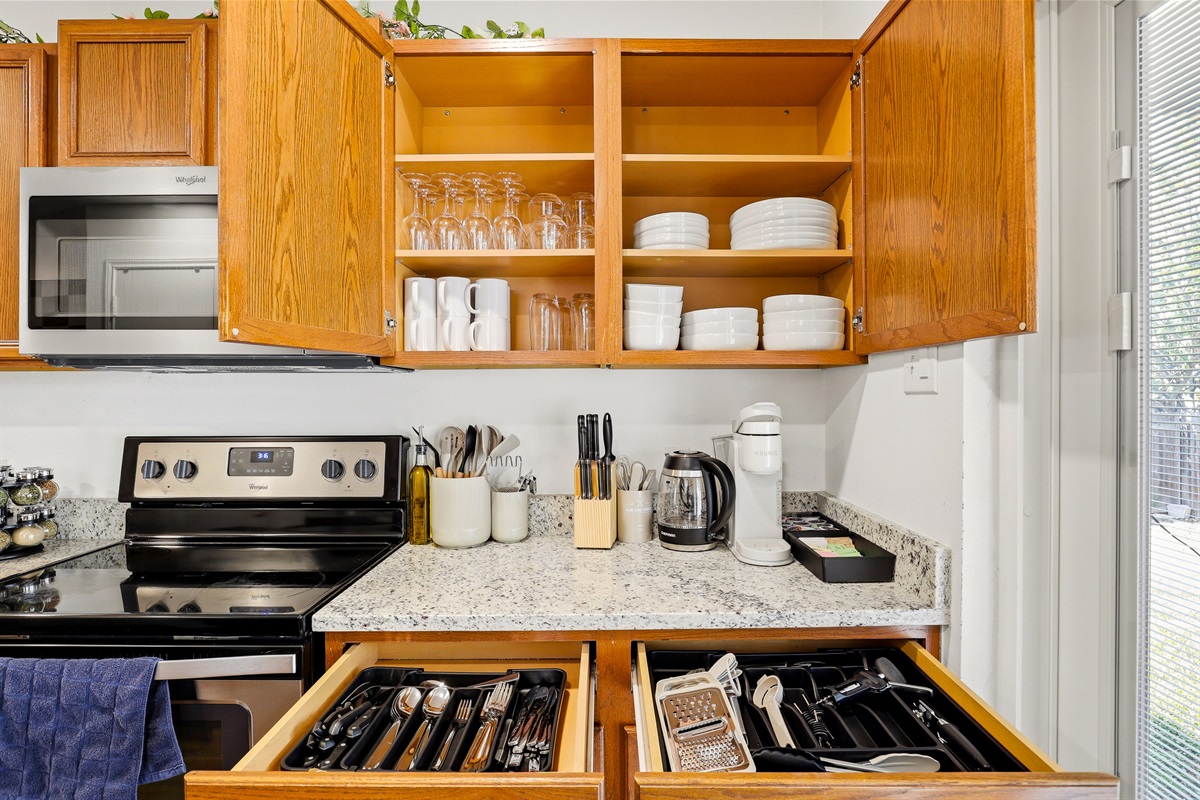 Fully stocked kitchen with wine glasses, plates, mugs, and full silverware drawers — plus electric stove, microwave, and Keurig coffee maker. No supply runs needed, just cook and enjoy.