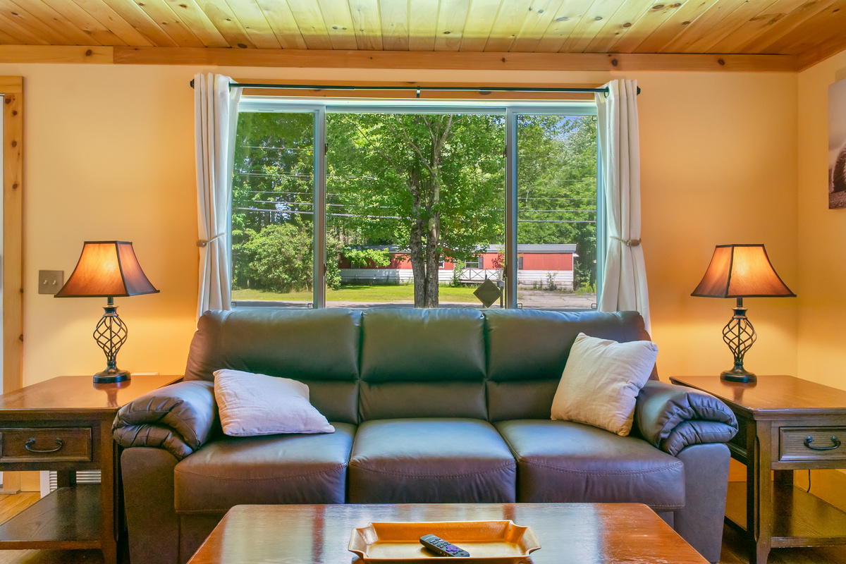 Relax on the spacious three-cushion couch positioned perfectly under a wide picture window. Flanked by matching wooden end tables with warm ambient lamps, this cozy living room setup features beautiful tongue-and-groove natural pine ceilings.