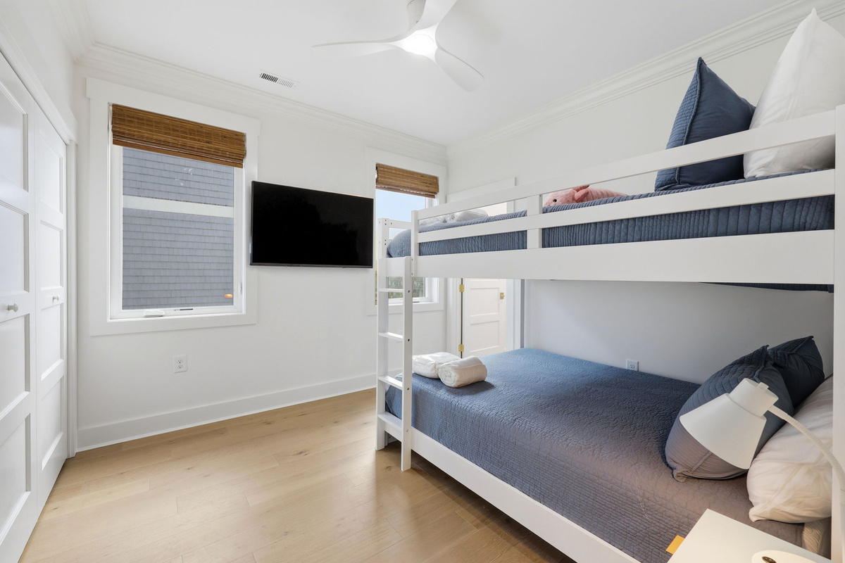 Queen-over-Queen bunk bedroom with coastal bedding, wall-mounted TV, and natural light