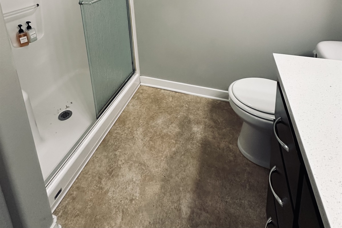 A bathroom featuring a clear glass shower with two soap dispensers on the wall, a white toilet, a countertop with a light-colored surface, and a neutral-colored floor.