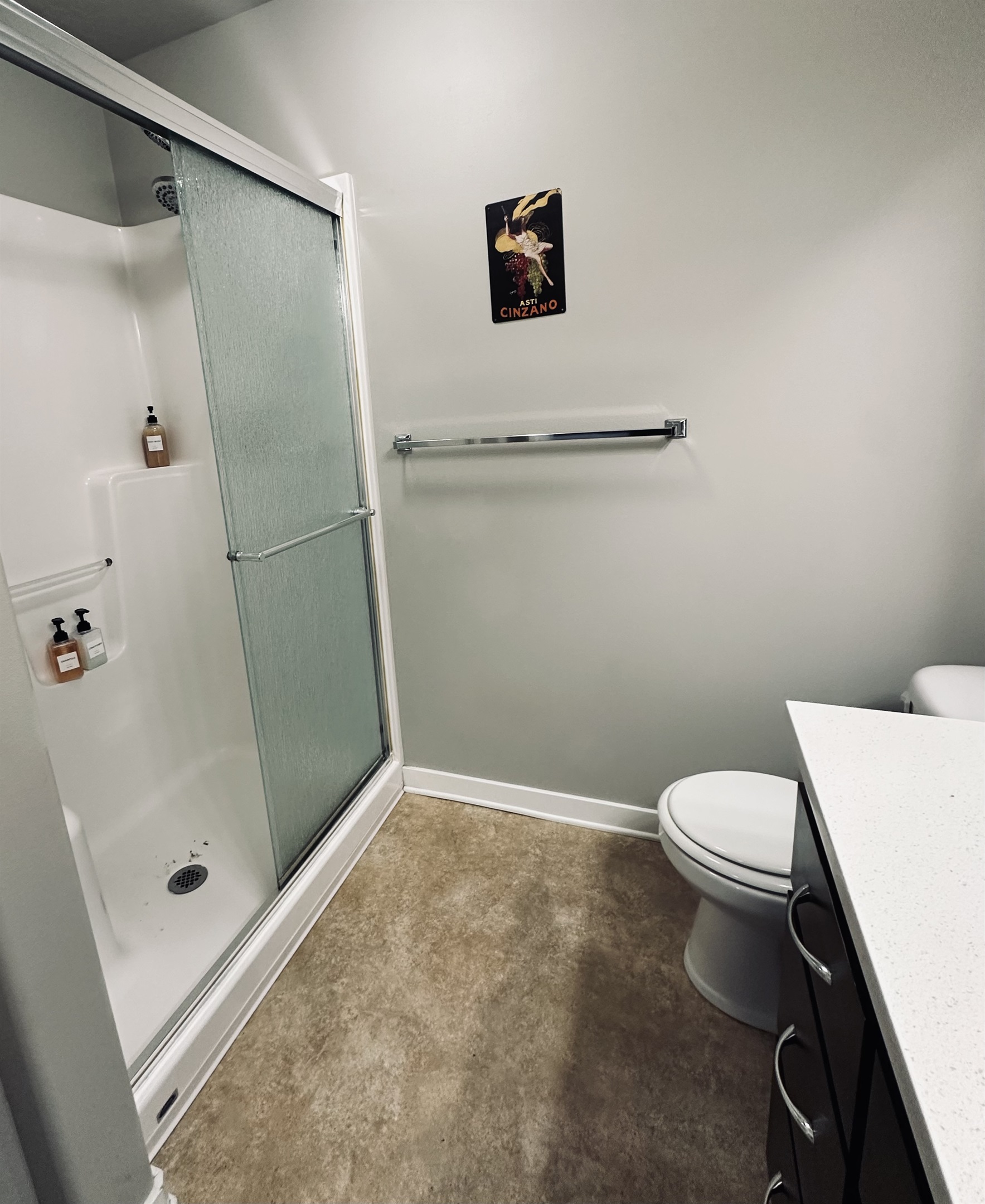 A small bathroom featuring a clear glass shower stall, a white toilet, a bathroom vanity with a light countertop, and a decorative poster hung on the wall.