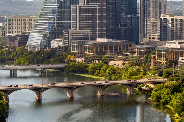 A panoramic view of a vibrant cityscape, showcasing a river with multiple bridges and lush green trees lining its banks. In the background, a stunning skyline of modern high-rise buildings and corporate offices dominates the view, suggesting a bustling urb