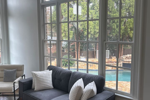 A serene living space framed by a tall arched window floods the room with natural light. A charcoal sofa with textured white pillows sits before a sparkling backyard pool and lush trees, blending modern comfort with elegant architectural detail.