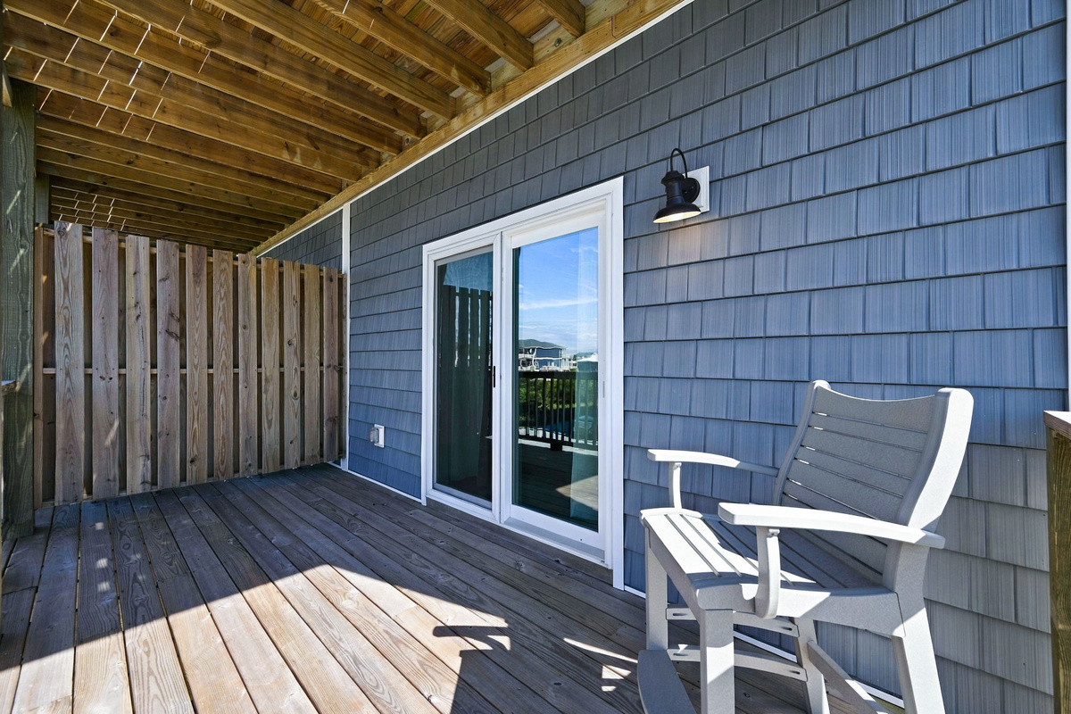 Covered private wooden deck area featuring coastal blue shaker siding, a comfortable bench seat, exterior sconce lighting, and sliding glass door access, enclosed with a vertical privacy wall.