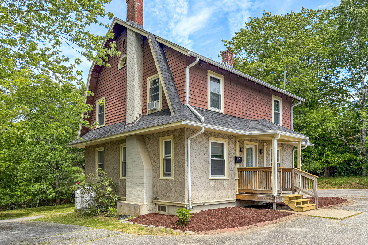This architectural view highlights the historic stone-pressed stucco siding, brick chimney, and gambrel roof. An upper window AC unit ensures climate control, verifying functional modern comfort at this private Downeast vacation home rental.