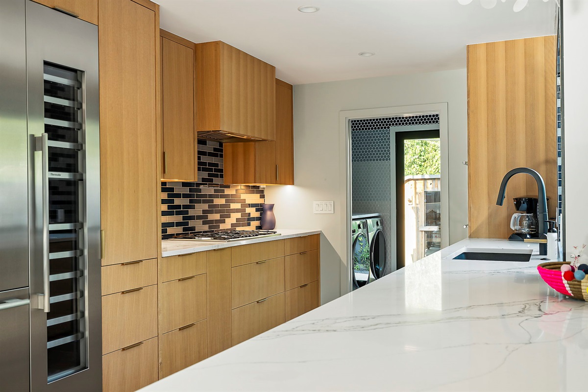 A sleek, modern kitchen featuring light wood cabinetry, a striking blue and black tile backsplash, and high-end stainless steel appliances, with a glimpse of a laundry area visible through a doorway at the back.