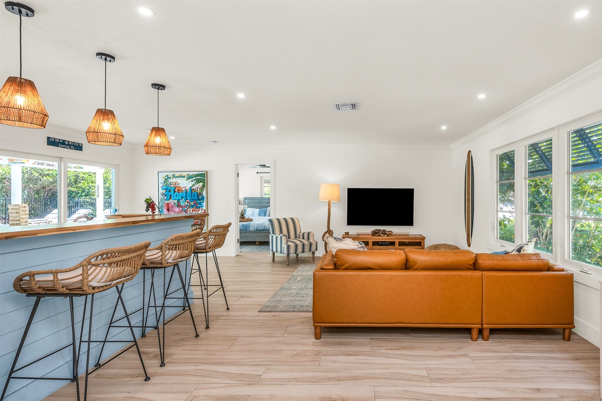 The bright and airy living room features a modern leather sectional, warm wood accents, and lush greenery views through large picture windows. Stylish touches like the surfboard décor and geometric coffee table create a relaxed coastal vibe.