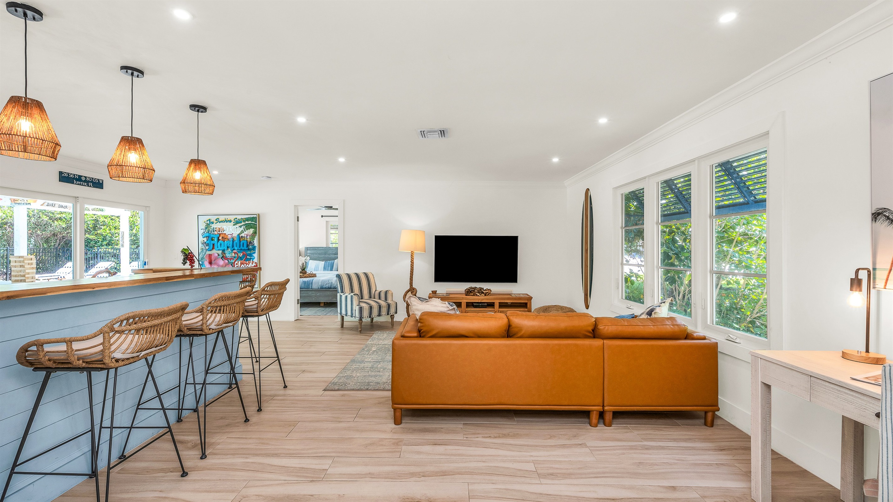 The bright and airy living room features a modern leather sectional, warm wood accents, and lush greenery views through large picture windows. Stylish touches like the surfboard décor and geometric coffee table create a relaxed coastal vibe.