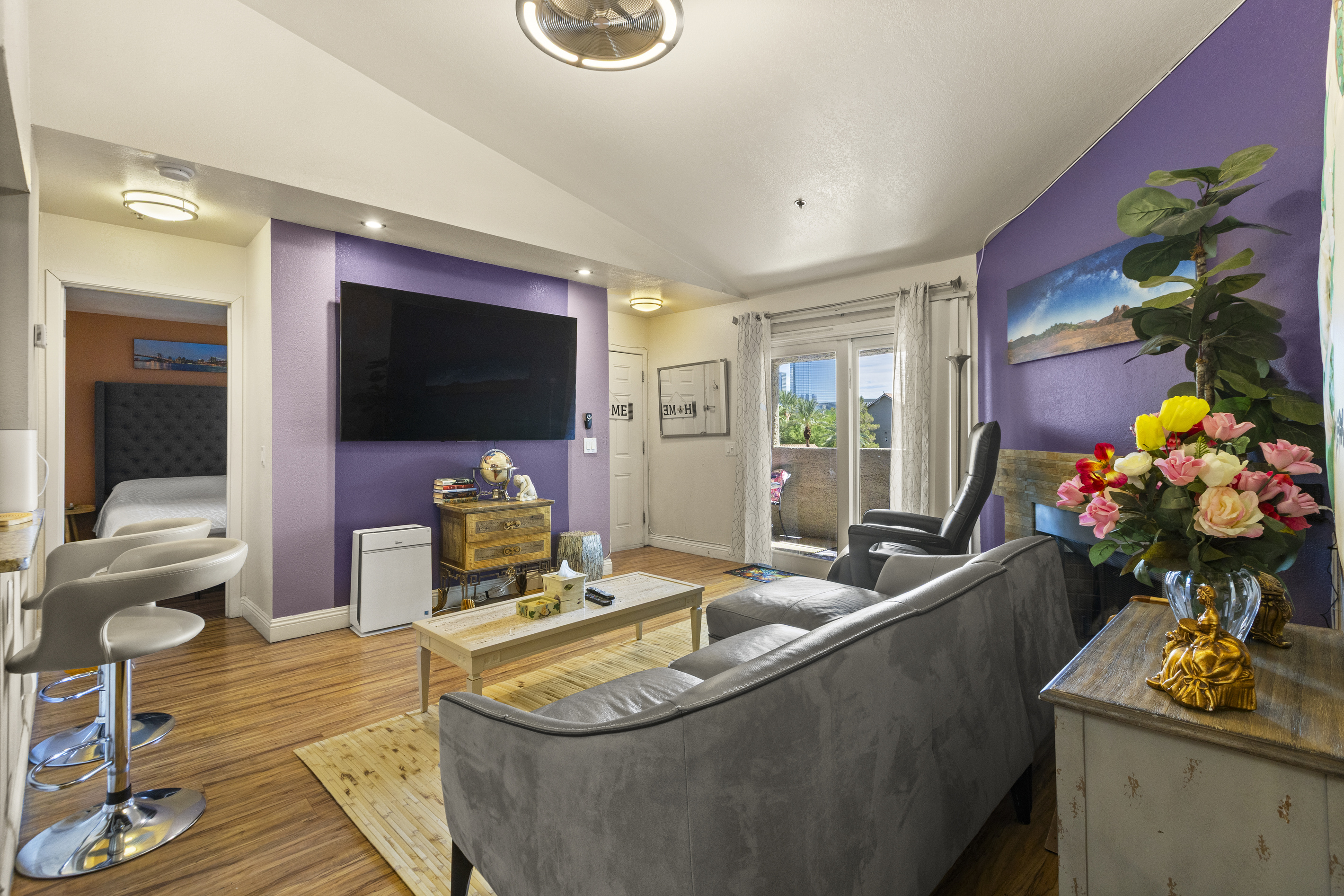 Living room with gray sofa seating, recliner, wall-mounted TV, balcony access, and a small bar counter nearby.
