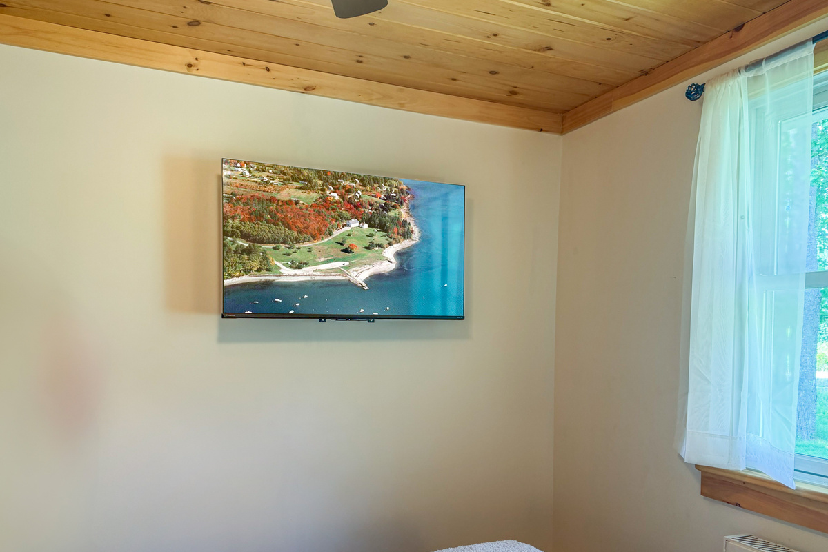 Enjoy seamless streaming on the large wall-mounted flat-screen TV in the guest bedroom. This straight-on angle captures the tongue-and-groove natural pine ceilings, clean neutral walls, and a bright window that fills the bedroom with daylight.