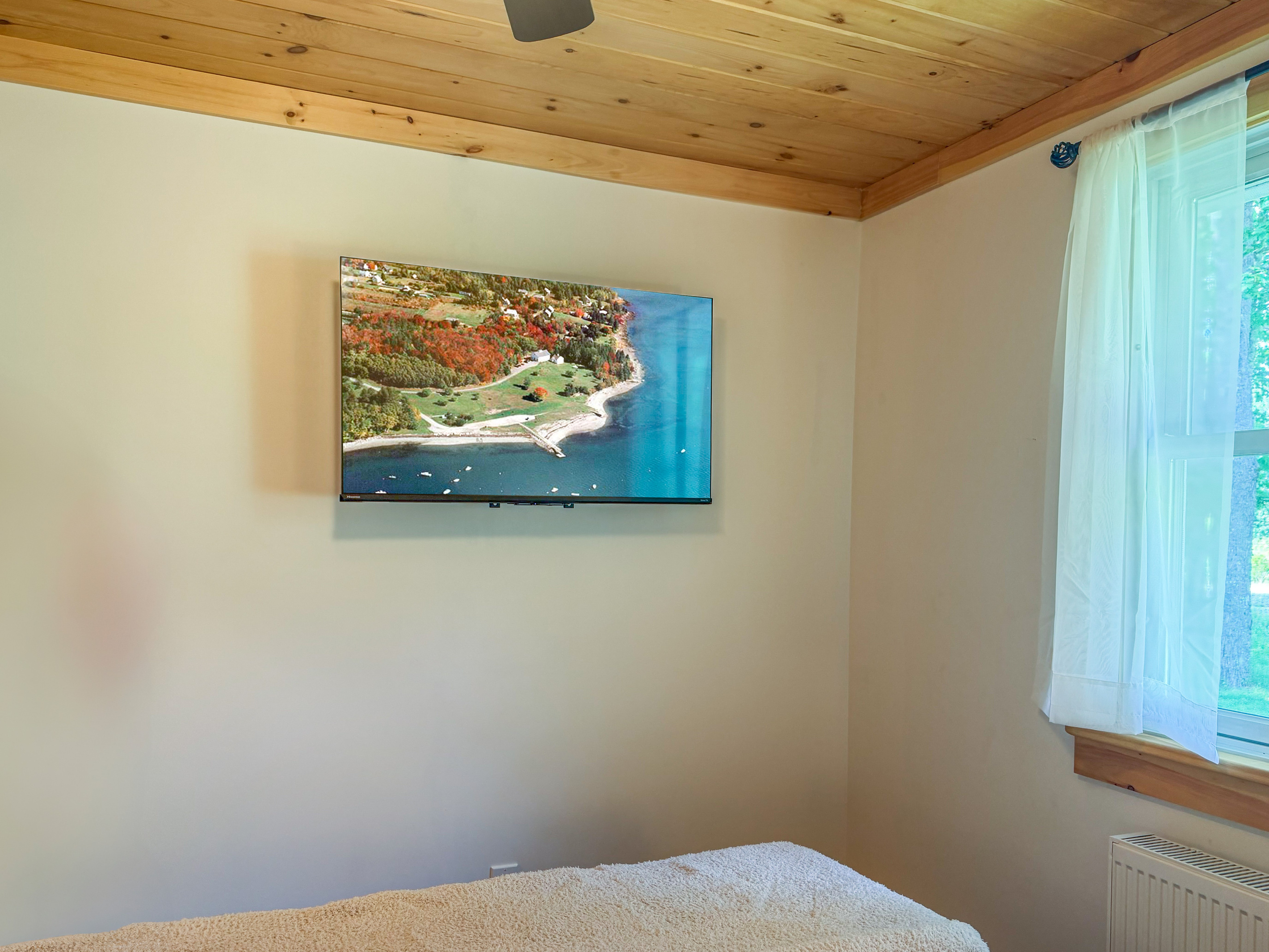 Enjoy seamless streaming on the large wall-mounted flat-screen TV in the guest bedroom. This straight-on angle captures the tongue-and-groove natural pine ceilings, clean neutral walls, and a bright window that fills the bedroom with daylight.