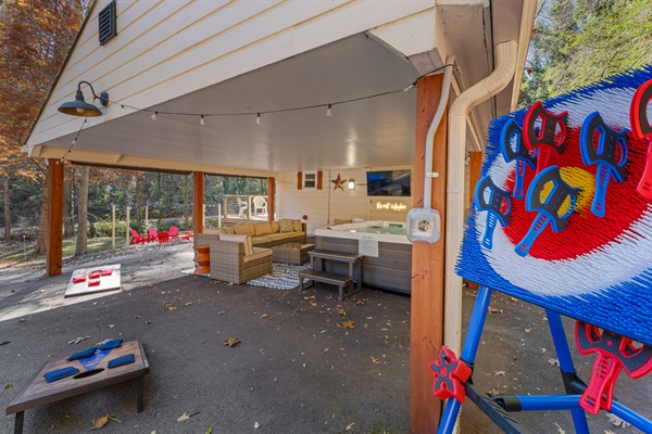 Covered outdoor hangout with hot tub, big-screen TV, lounge seating, and lawn games — perfect for enjoying the fresh air and forest views.