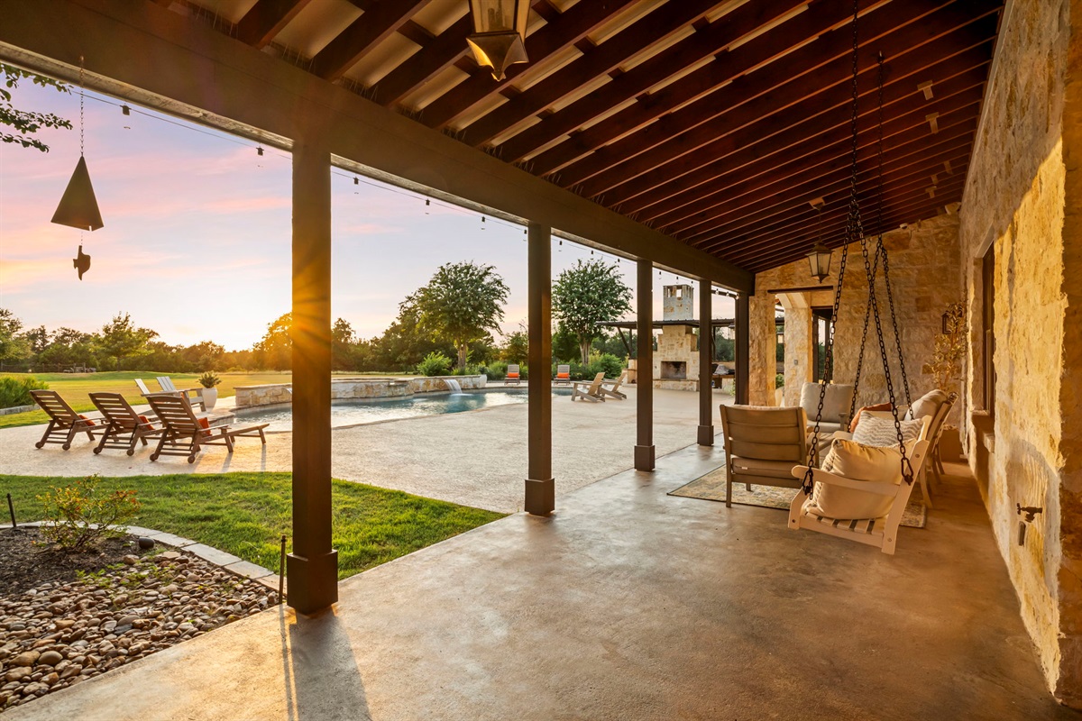 A tranquil evening on a spacious covered patio overlooking a sparkling pool and a lush green lawn, bathed in the warm glow of a setting sun.
