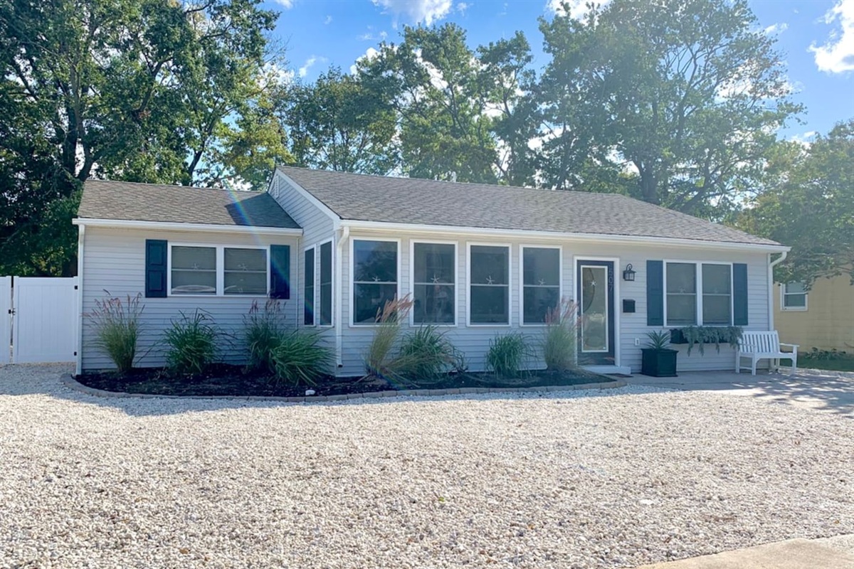 Welcome to your getaway! This lovely ranch-style home features light gray siding, navy shutters, and a bright sunroom. The exterior is complete with a white stone and manicured greenery, offering great curb appeal and a peaceful setting.