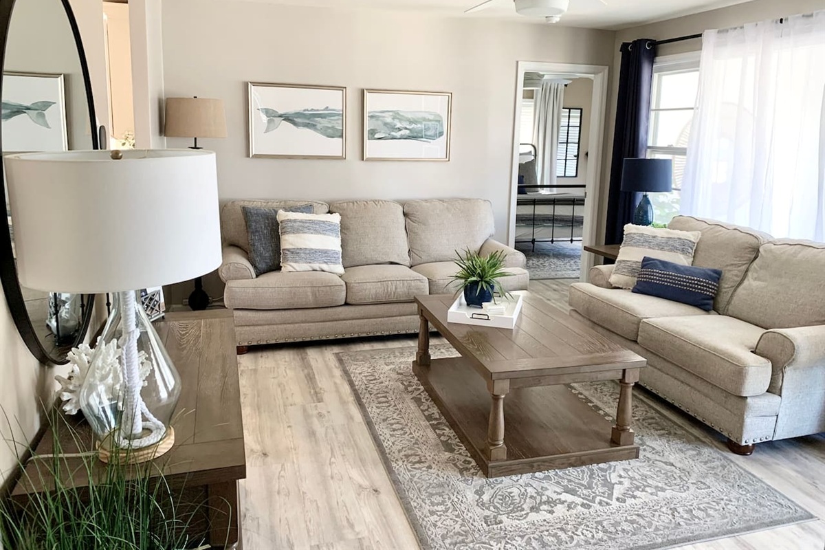 Relax in our coastal living room featuring comfy beige sofas, nautical pillows, and whale art. This bright, airy space with a rustic coffee table and patterned rug is perfect for family time after the beach.