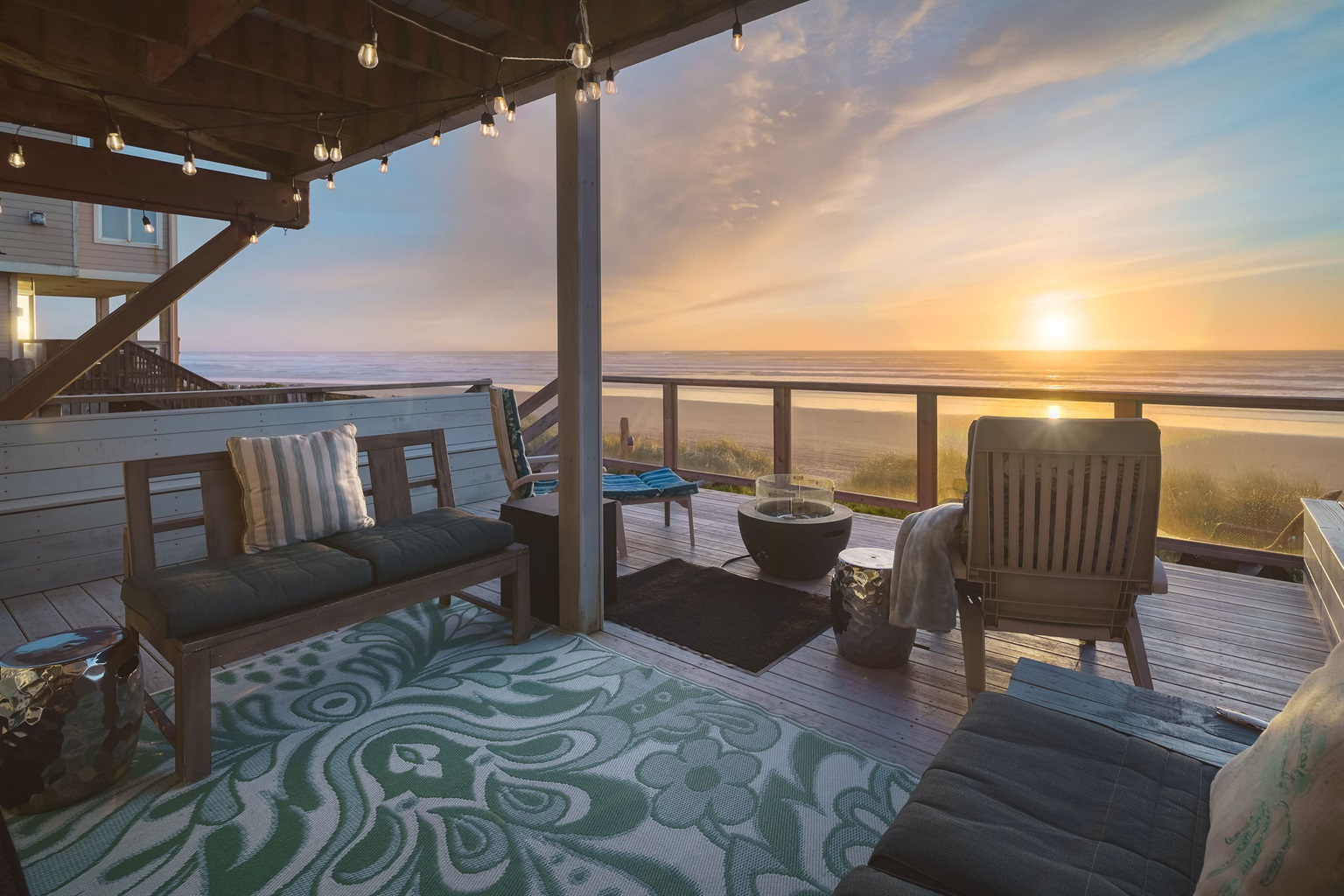 With beachfront scene viewed from a cozy, well-furnished wooden deck. The deck is adorned with an array of comfortable outdoor furniture, including a fire pit and string lights that invite relaxation. 
