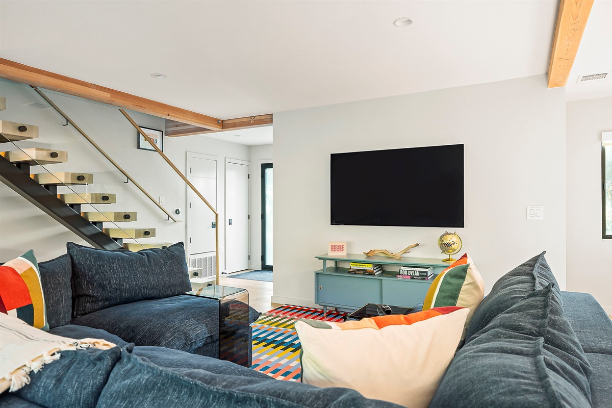A stylish and vibrant living area with a large sectional sofa, a wall-mounted television above a colorful console, and a modern floating staircase with a glass railing.