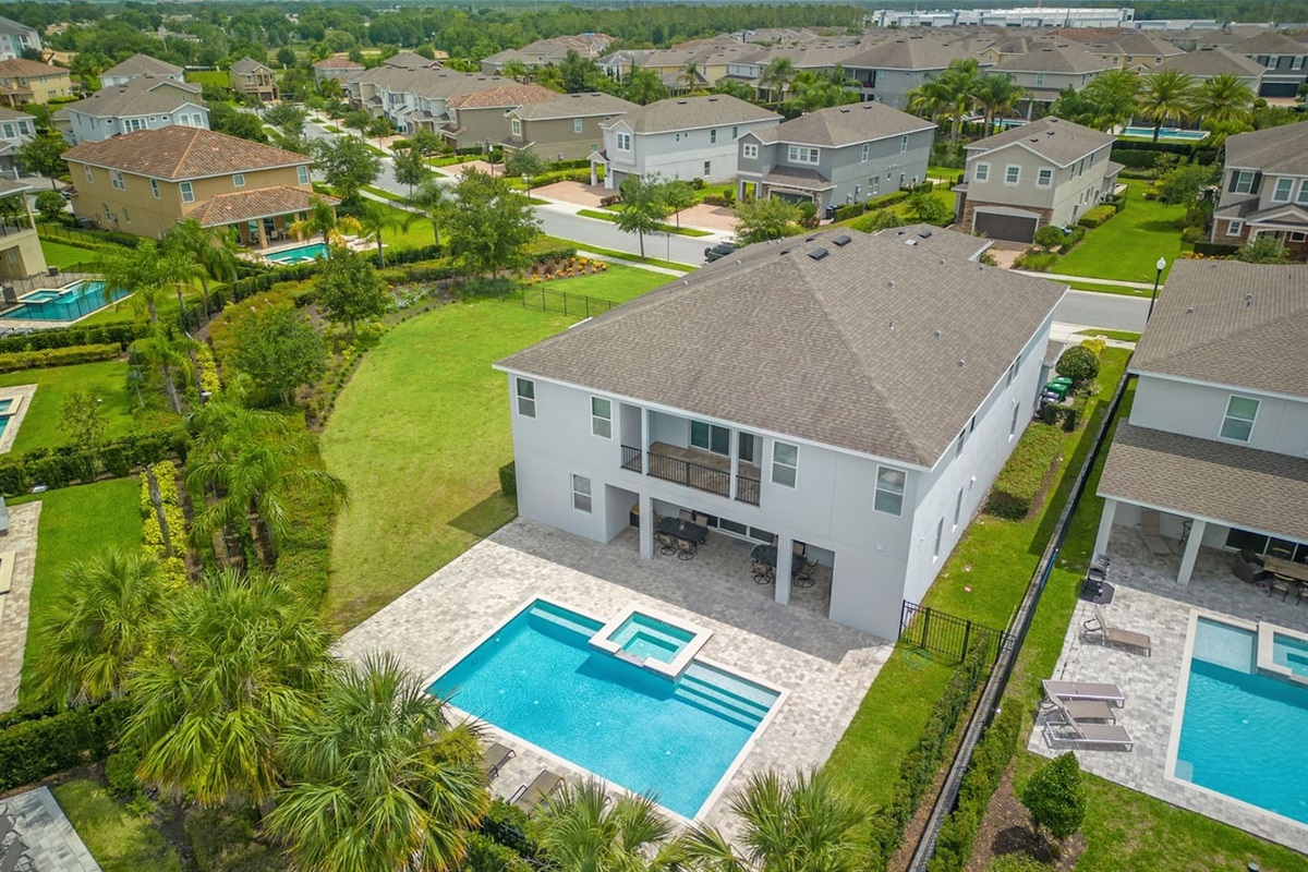 This aerial view showcases a spacious backyard with a sparkling pool, hot tub, and a cozy outdoor patio area. The home is nestled in a beautiful, serene neighborhood with plenty of greenery, offering a relaxing retreat for your next stay.