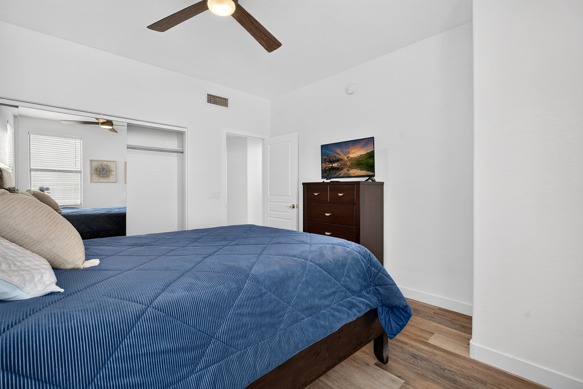 Queen bedroom with ceiling fan, TV, closet storage, side table, and natural light from the corner windows.