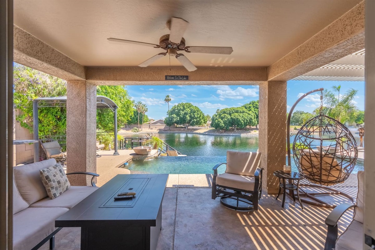 The covered patio is your outdoor living room — a plush sofa, fire table, ceiling fan, and that swinging egg chair frame a view of the pool and lake that never gets old.