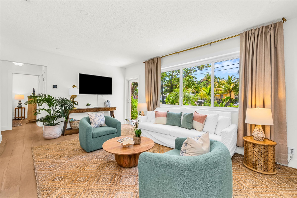 Elegant, sun-filled living room featuring designer coastal décor, plush swivel chairs, a chic white sofa, and custom wood accents, with tropical views and a seamless flow into the gourmet kitchen—perfect for luxurious relaxation and entertaining.