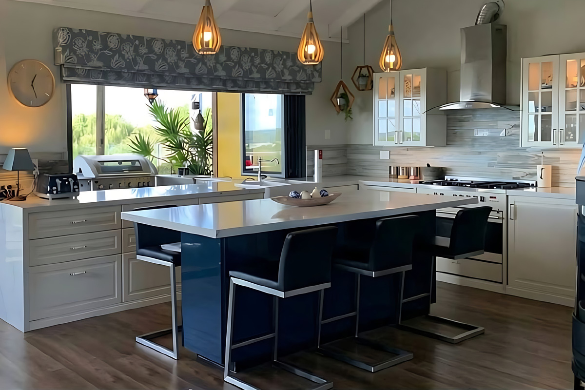 An island bench with four stools sits at the centre of the kitchen, with a freestanding gas cooker, glass-front cabinets and a window over the sink that opens out to the outdoor BBQ.
