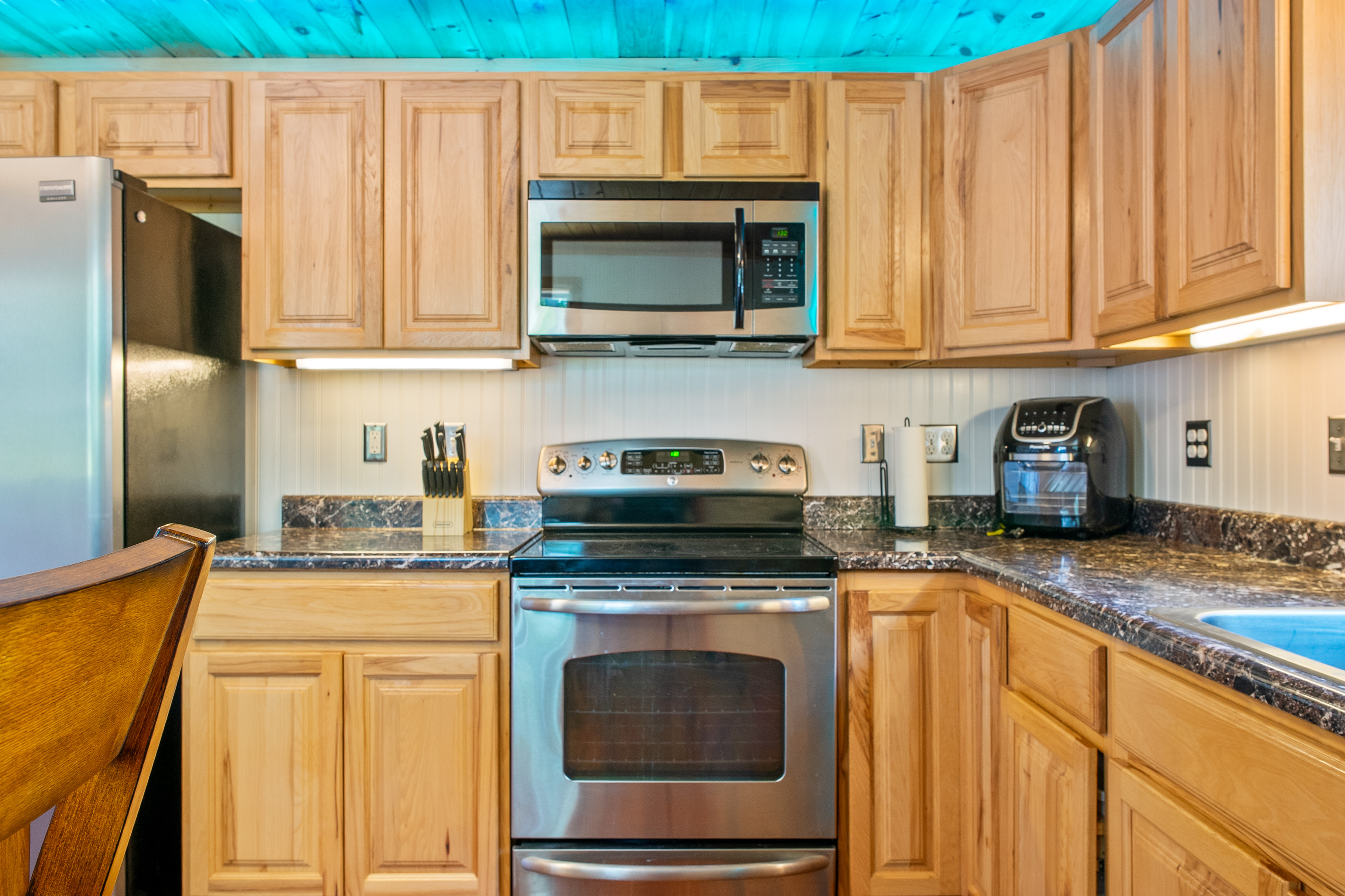 Prepare group feasts using the stainless steel electric range and oven with a matching over-the-range microwave. The L-shaped kitchen counter setup features a knife block, under-cabinet lighting, and a countertop air fryer for easy meals.
