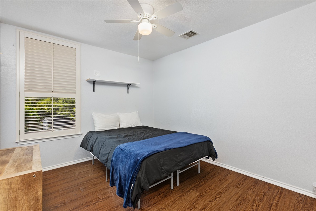 Queen bedroom with soft navy bedding, ceiling fan, window blinds, and clothing storage. Hotel-quality linens and extra pillows included. Quiet and private with hardwood floors in this 5BR San Antonio home.