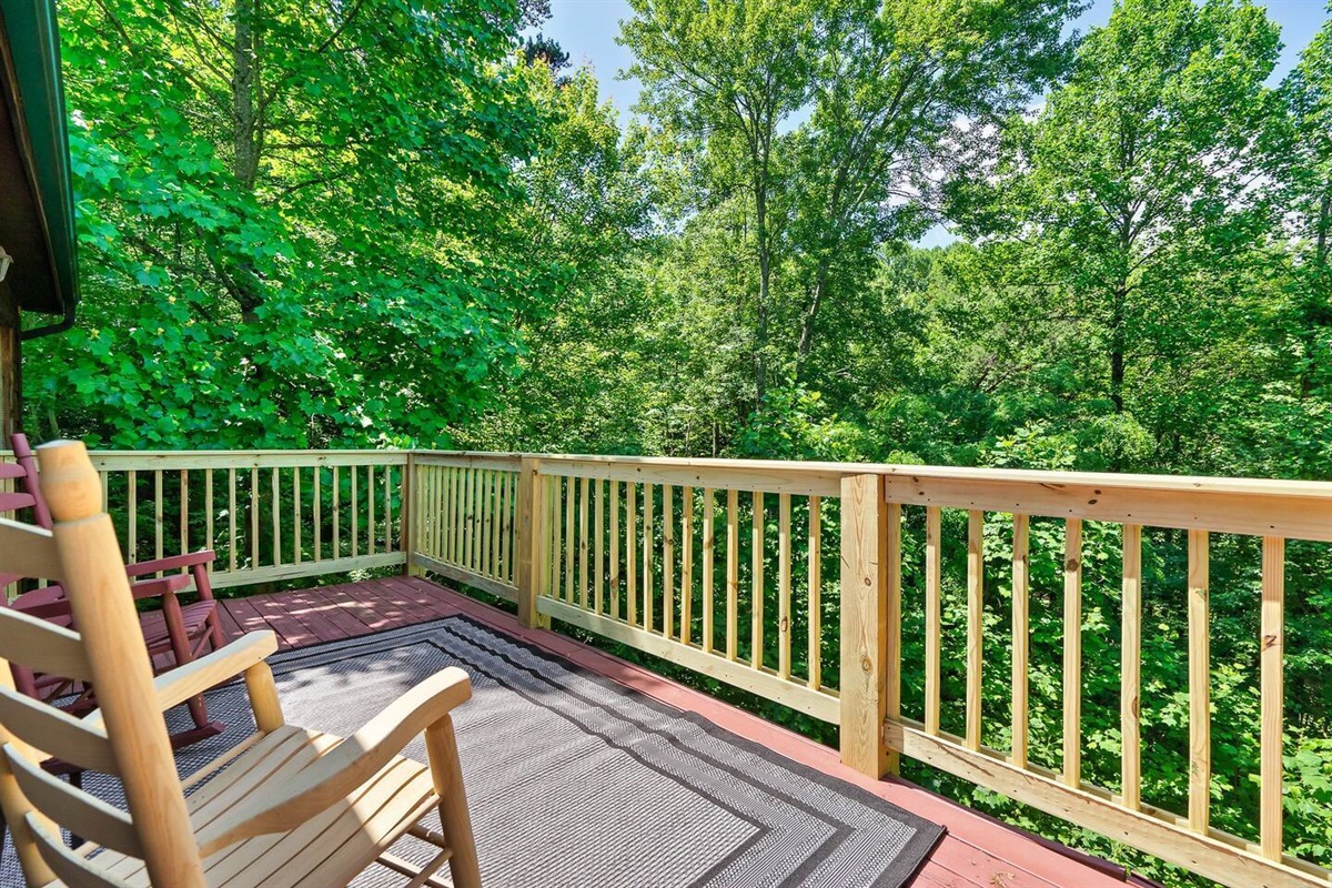This spacious outdoor deck offers a tranquil retreat with comfortable rocking chairs and a wooden railing, providing panoramic views of the vibrant green treetops.