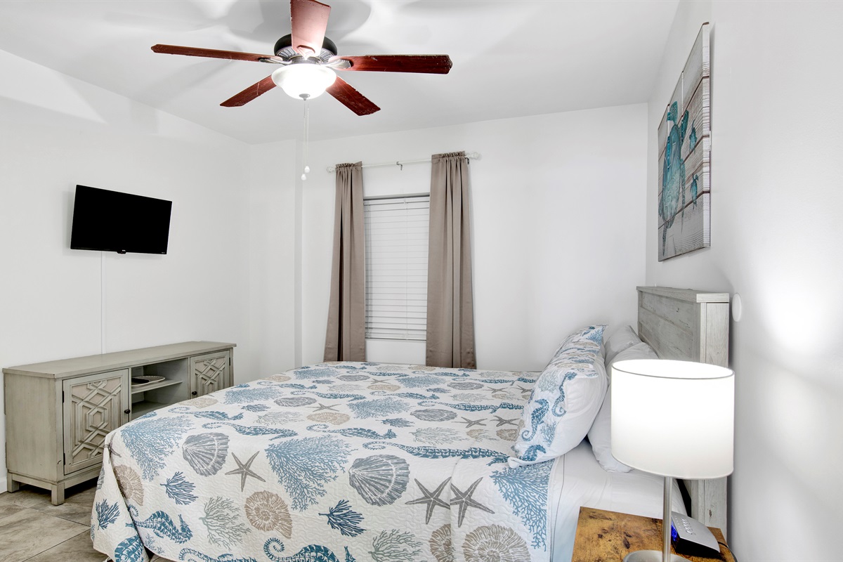 Bedroom with king bed, wall mounted TV, ceiling fan, dresser and ensuite bathroom