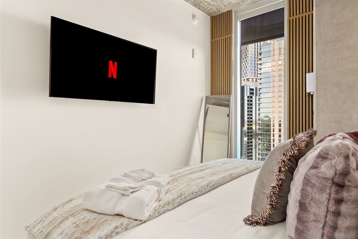 A comfortable bedroom featuring a wall-mounted TV with Netflix on the screen, a large window offering a view of a cityscape, and a full-length mirror, creating a cozy yet modern and stylish retreat.