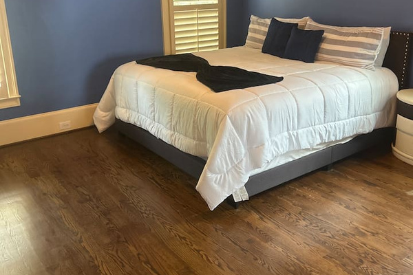 Bright, cozy bedroom with a comfortable queen bed, crisp white linens, and plush pillows. Calming blue walls, hardwood floors, and natural light from shuttered windows create a relaxing retreat. Includes nightstand and ample space to unwind.