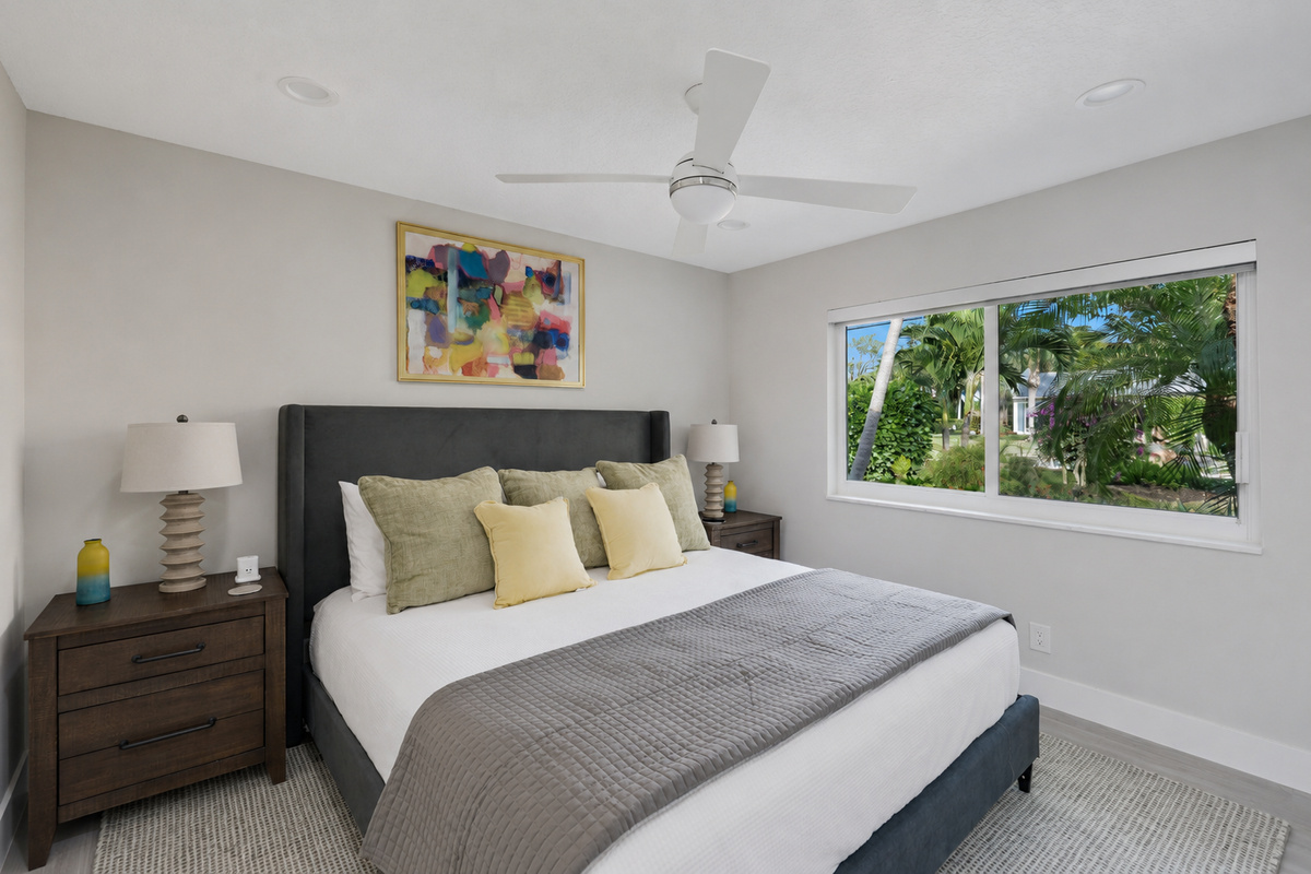 Rest in a bright king bedroom with soft bedding, tropical window views, a ceiling fan, and a calm place to recharge.
