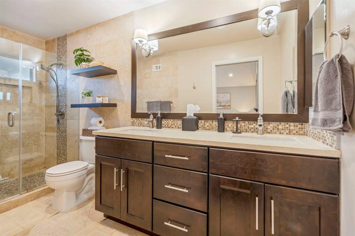 A modern primary bathroom featuring a spacious walk-in shower with a handheld shower head, dual sinks with sleek faucets, a large mirror stretching across the vanity, and ample overhead and wall-mounted lighting for a bright, functional space.