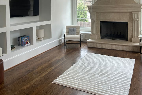 Airy living room with soaring vaulted ceilings, exposed wood beams, and a tall arched window flooding the space with light. A stone fireplace anchors the room, balanced by built-in shelving, warm hardwood floors, and a soft neutral rug.