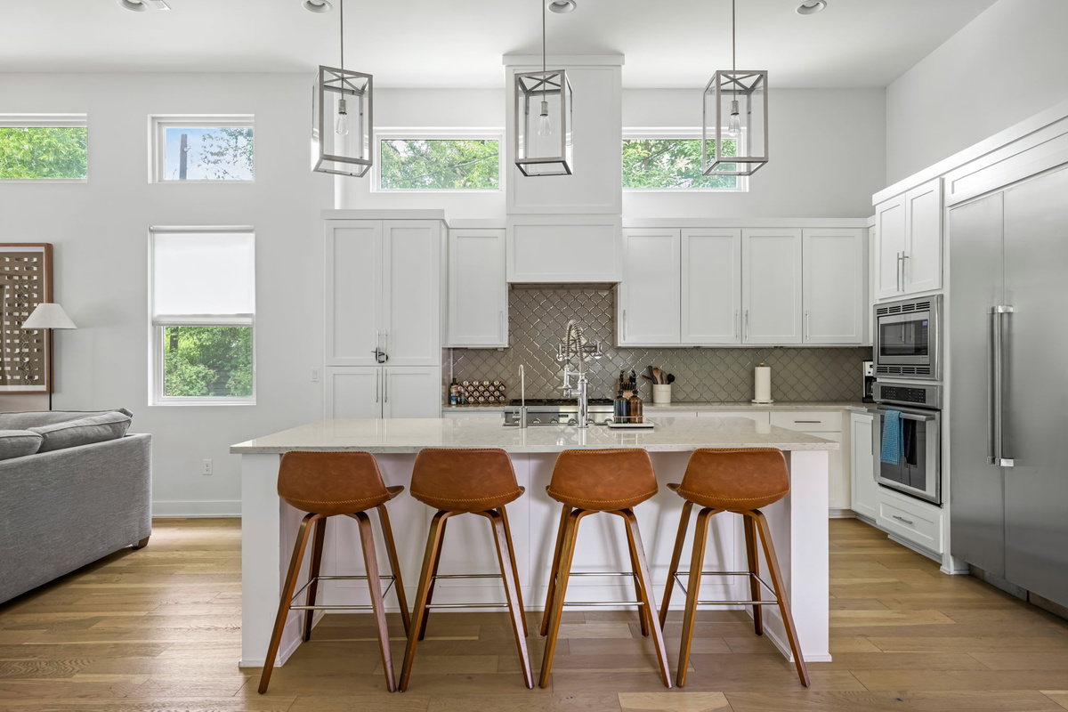 Expansive quartz island with warm leather stools and stainless appliances, framed by tall windows and abundant natural light.
