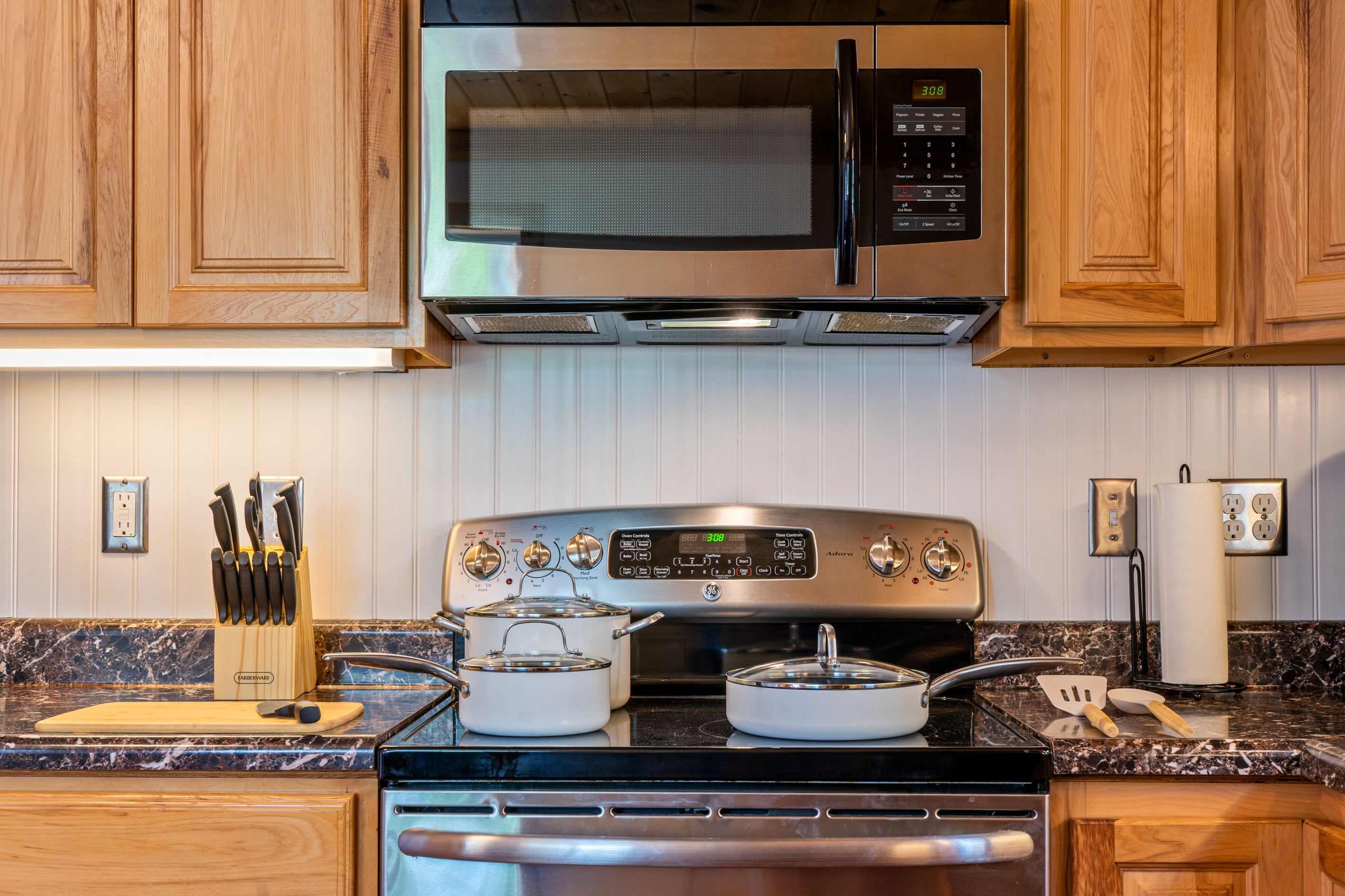 Cook effortlessly utilizing the stocked ceramic-coated nonstick pots and pans set arranged on the glass cooktop. The station features an electric range, an over-the-range microwave, a wood cutting board, and a Farberware knife block set.