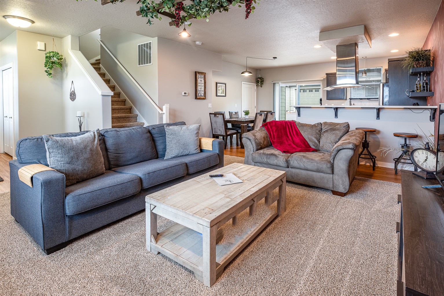 A cozy living room featuring a blue sofa and a brown leather couch with a red throw blanket, a rustic wooden coffee table in the center, and a dining area with a set of dark wooden chairs in the background, along with a staircase leading to the upper level.