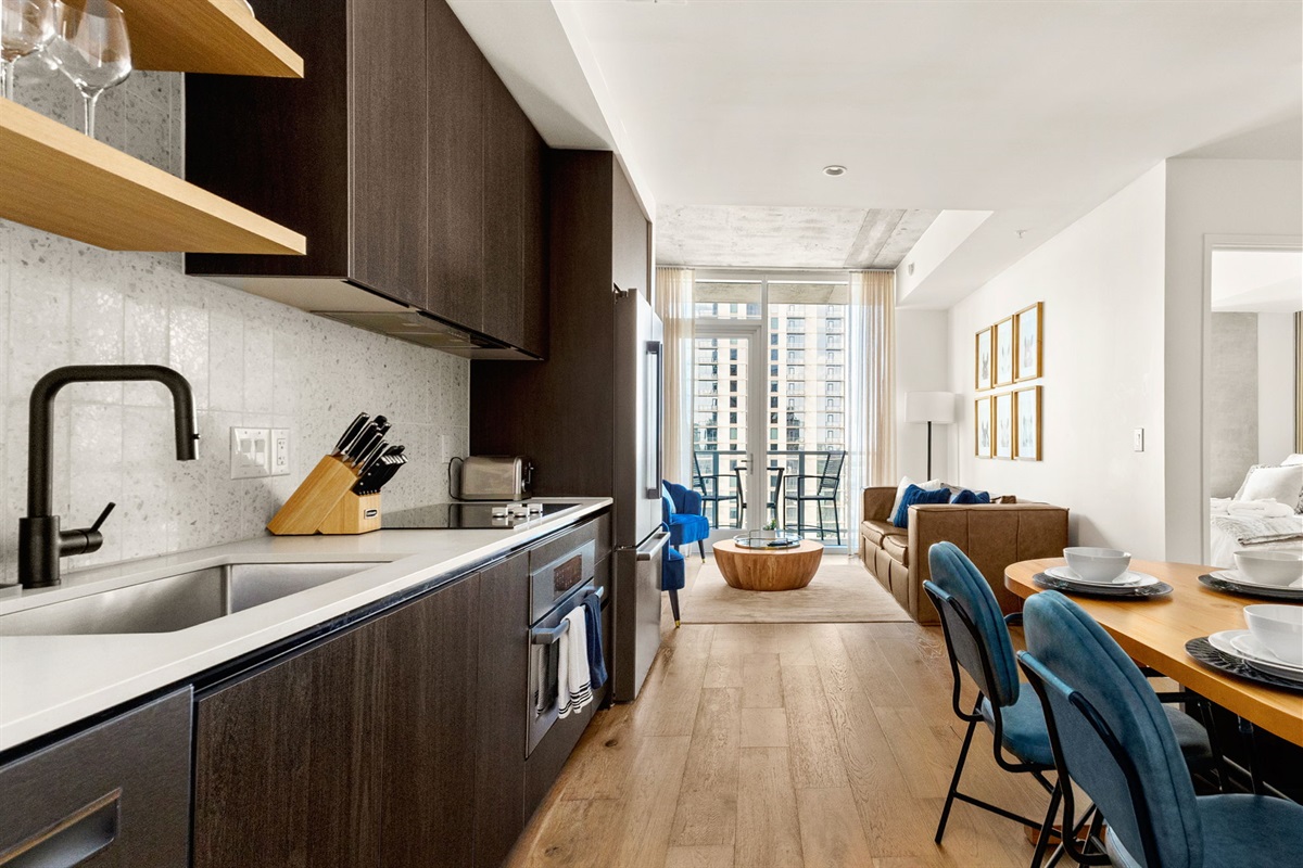 A contemporary open-concept space featuring a modern kitchen with dark wood cabinetry and a white speckled backsplash. The area flows into a dining nook with a wooden table and stylish blue chairs, and then into a bright living room with a comfortable sofa