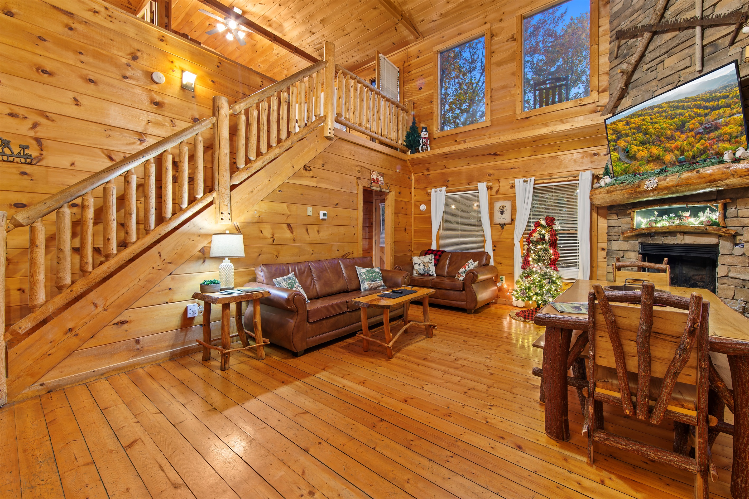A warm and rustic living space with polished wood floors, a staircase leading to a loft, and an adjacent kitchen with black appliances and dining. The cozy vibe is enhanced by leather couches and charming decor.