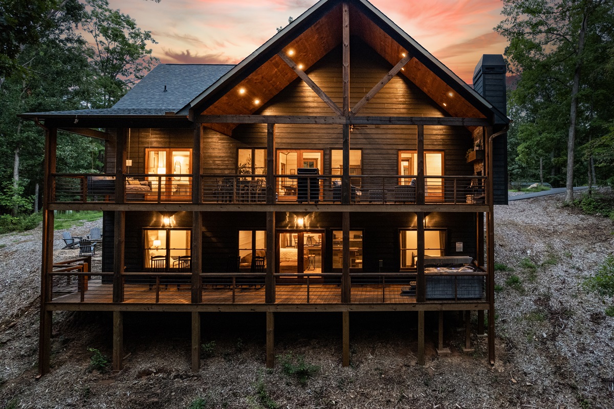 Rear exterior of the cabin showing layout of decks with hot tub, and rockers on lower level and fireplace, grill, eating and sitting areas and hanging swing on upper level.