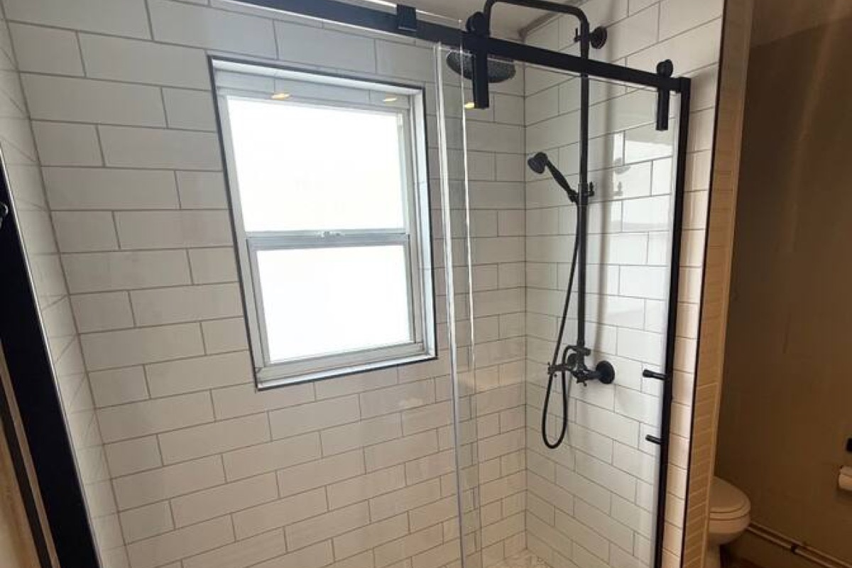 Newly upgraded bright, modern walk-in shower w/ white subway tile and classic hex tile floor. Features black rain shower head, handheld sprayer, and sliding glass barn-style door. Natural light from the frosted window keeps the space airy and private