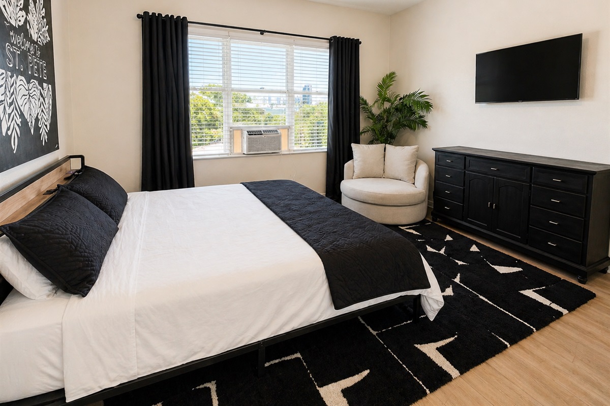 Stylish and cozy bedroom featuring a comfortable queen bed, soft linens, and modern décor. Enjoy natural light, a wall-mounted TV, and a relaxing reading chair—perfect for unwinding after a long day.