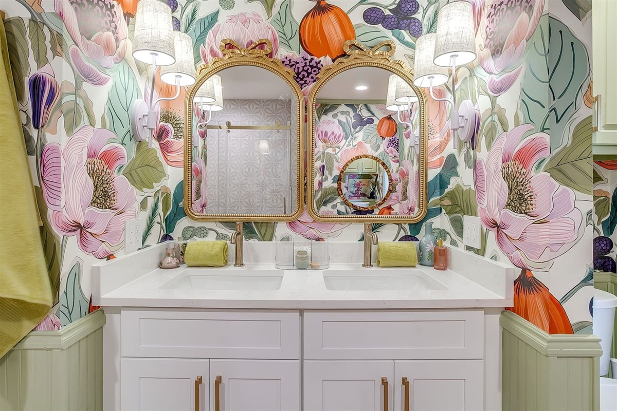 Rear bath with double vanities, statement wallpaper, gold fixtures, and classic chair railing — designed so everyone has space to get ready in style.